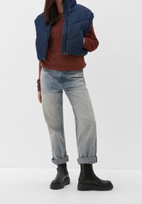 Gepolsterte blaue Weste mit hohem Kragen, bordeauxroter, strukturierter Pullover, hellblaue Jeans, an den Bündchen hochgekrempelt, und schwarze Stiefeletten mit dicken Sohlen.