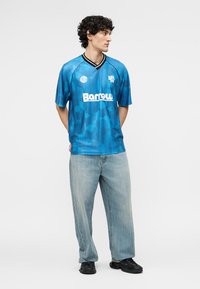 Νέος άνδρας που στέκεται με τα χέρια πίσω από την πλάτη, φοράει μπλε αθλητική φανέλα με σχέδιο και τη λέξη «Barrow» και ανοιχτόχρωμα χαλαρά τζιν.