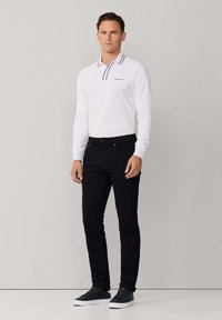 Polo blanco de manga larga con detalles y logo en azul marino, combinado con jeans negros ajustados y zapatillas gris oscuro. Diseño sencillo y moderno.