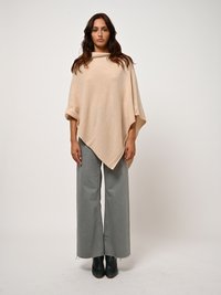 Beige Strick-Poncho mit hohem Kragen, nach unten gerichtet, kombiniert mit weit geschnittenen hellgrauen Hosen und dunkelgrünen Absatzstiefeln.