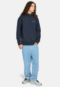 Marineblauwe fleece pullover met een rits-kraag, gecombineerd met lichtblauwe geblokte jeans en grijze sneakers.