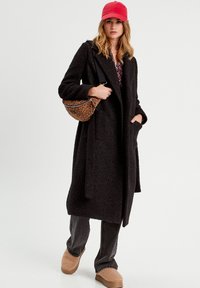 Black textured long coat with pockets, paired with dark jeans and beige platform clogs. Accessorized with a leopard print bag and red cap.