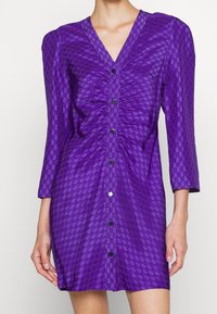 Lila klänning med V-ringning, trekvartslånga ärmar, rynkade detaljer och knäppning. Tillverkad av strukturerad tyg med mönstrade detaljer.