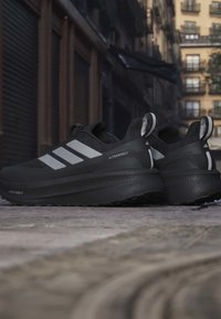 Sorte sneakers med et strømlinet design, der har tre reflekterende hvide striber, en tekstureret overdel og en tyk gummisål til stødabsorbering.