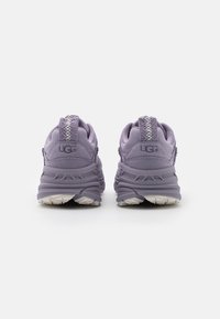 Violet urheilukengät, joissa on teksturoitu pinta, pehmustettu kaulus ja valkoinen kumipohja. Brändin logo on kohokuvioitu kantapäässä.