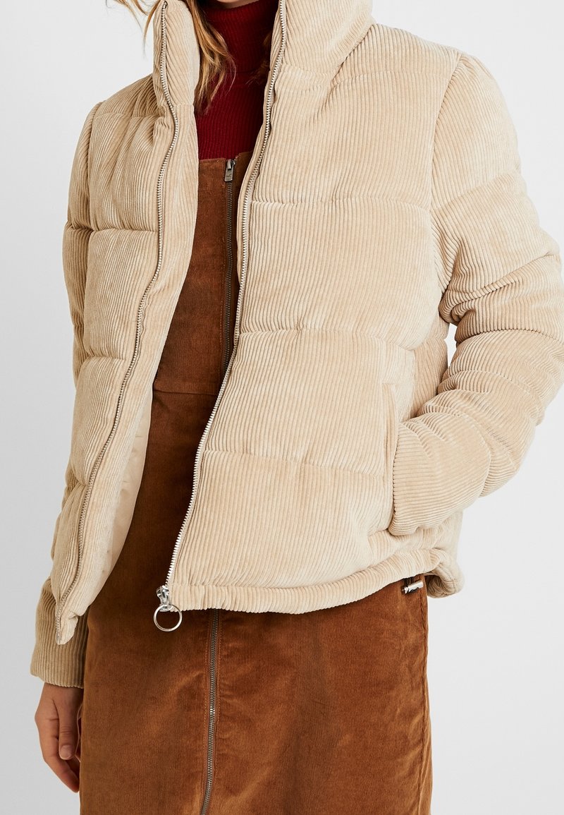 Beige corduroy pufferjakke med høj krave, sidelommer og lynlås foran. Båret over en brun corduroy kjole.
