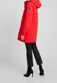 Rode gewatteerde parka met een capuchon, bontafwerking, logo-opdruk op de mouw en voortassen. Combinatie met zwarte leren broeken en zwarte leren laarzen met hak.