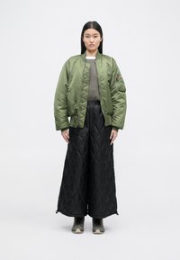 Alyvuogių žalios spalvos bombonešis su siuvinėta tekstūra, derinamas su juodomis plačiomis kelnių kelnes. Tamsūs batai užbaigia įvaizdį.