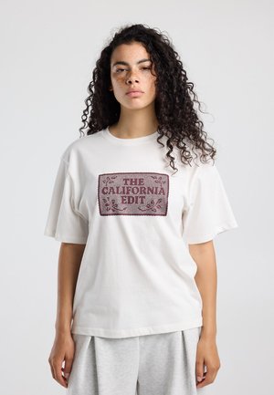 Jeune femme aux cheveux bouclés et aux taches de rousseur porte un t-shirt blanc oversize avec le texte "The California Edit" et un pantalon gris clair, debout devant un fond uni.