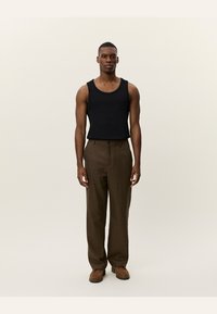 Black sleeveless tank top paired with brown checkered trousers and brown loafers, standing against a neutral background.