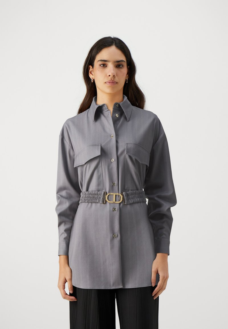 Chemise grise à manches longues avec des rayures, deux poches poitrine, fermeture à boutons et ceinture élastique avec une boucle dorée. Texture de tissu lisse.