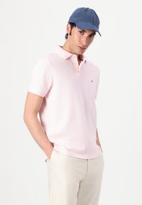 Roze polo shirt met een kraag en korte mouwen, gecombineerd met crèmekleurige broeken. Een blauwe pet ligt op het hoofd, met een klein logo.