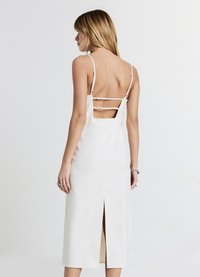 Eine Frau trägt ein ärmelloses, rückenfreies weißes Kleid mit dünnen Trägern und zwei horizontalen Rückenträgern. Sie steht vor einem schlichten weißen Hintergrund.