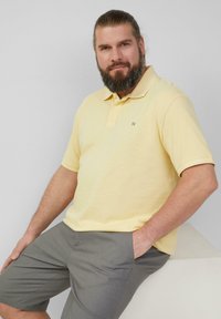 Gelbes Poloshirt mit strukturierter Oberfläche, zwei Knöpfen am Kragen und einem kleinen Logo auf der linken Brust, kombiniert mit grauen Shorts.