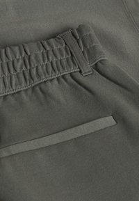 Gros plan sur la taille de pantalons vert olive avec un band élastique et une passante de ceinture au-dessus d'une poche horizontale.