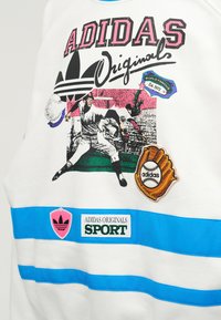 Παίκτης μπέιζμπολ ρίχνει με λογότυπο Adidas, ετικέτα γαντιού, κείμενο "Adidas Originals" και μπλε ρίγες σε λευκό ύφασμα.