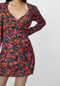 Vestido floral con escote en V profundo, con un fondo oscuro y patrones coloridos en rosa, naranja y verde. Mangas largas con detalles de fruncido.