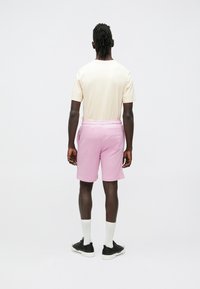 Világos bézs rövid ujjú ing és rózsaszín rövidnadrág rugalmas derékpánttal. A modell fekete cipőt és fehér zoknit visel, hátrafordulva a kamerától.