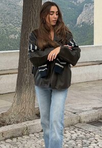 Jonge vrouw met lang haar, gekleed in een zwarte leren jas, lichtblauwe spijkerbroek en een gestreepte trui over haar schouders gedrapeerd, staand buiten.