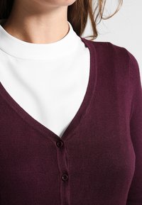 Bordowy kardigan z dekoltem w serek wykonany z gładkiej dzianiny, zapięcie na małe ciemne guziki i lekko dopasowany krój.
