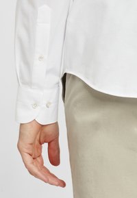 Witte button-up shirt met ronde manchet en twee knopen, gecombineerd met lichtbeige broek. Gladde stof met een minimalistisch ontwerp.