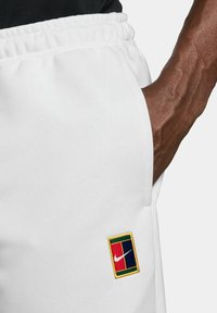 White athletic shorts with an elastic waistband, featuring a multi-colored embroidered Nike logo patch on the left pocket.