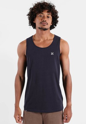 Homme aux cheveux bouclés portant un débardeur noir sans manches et un pantalon marron, se tenant contre un fond blanc uni.