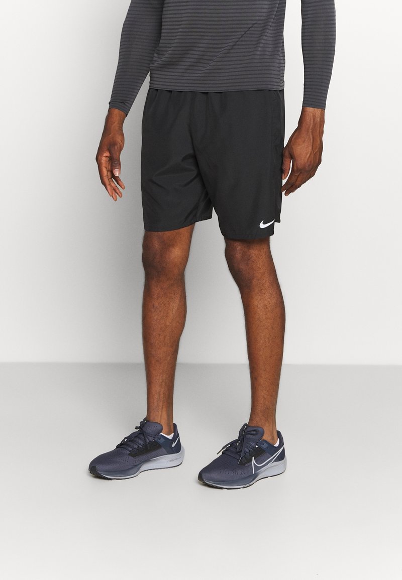 Homme portant un short Nike noir, une chemise à manches longues grise et des chaussures de course Nike gris foncé, debout sur un sol gris.
