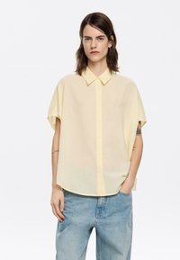 Yellow short-sleeved shirt with a collared neckline and button closure, made of lightweight material featuring a subtle textured pattern.
