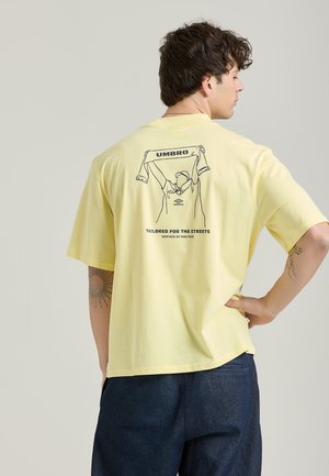 En gul T-shirt med kort ärm har en rygggrafik av en figur som håller en scarf, med "UMBRO" ovanför och texten "TAILORED FOR THE STREETS" nedanför.