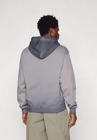 Siva ombre dukserica s podesivom kapuljačom, rebrastim manžetama i donjim rubom, izrađena od mekanog materijala, s opuštenim krojem i jednostavnim dizajnom.