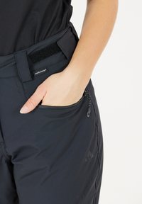 Zwarte broek van flexibel materiaal, met een klittenbandsluiting, een zijritszak en een klein logo op de dij.