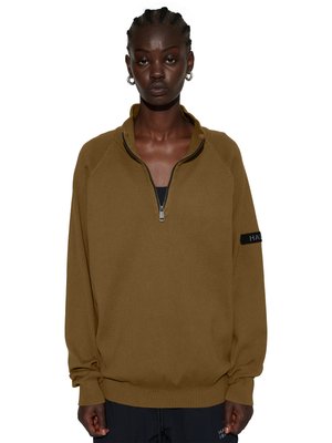 Osoba o ciemnej karnacji nosząca brązowy sweter z zamkiem do ćwiartki i czarną koszulkę pod spodem oraz srebrne kolczyki kółka, stojąca na białym tle.