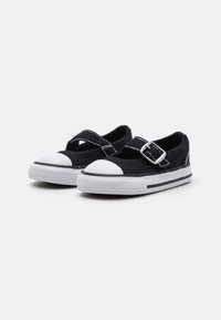 Zapatillas de lona negras con puntera y suela de goma blanca, que cuentan con una correa cruzada con una hebilla plateada y costuras blancas en contraste.
