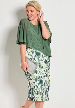 Lächelnde Frau in grünem Blumenkleid und Umhang, die eine silberne Clutch hält und vor einer schlichten hellen Wand posiert.