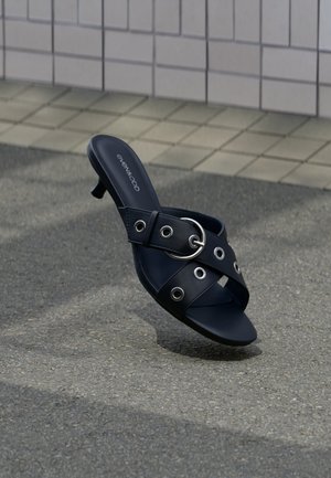 Czarna sandałka na obcasie z krzyżującymi się paskami ozdobionymi srebrnymi oczkami i okrągłą klamrą, umieszczona na teksturowanym chodniku obok ściany wyłożonej płytkami.