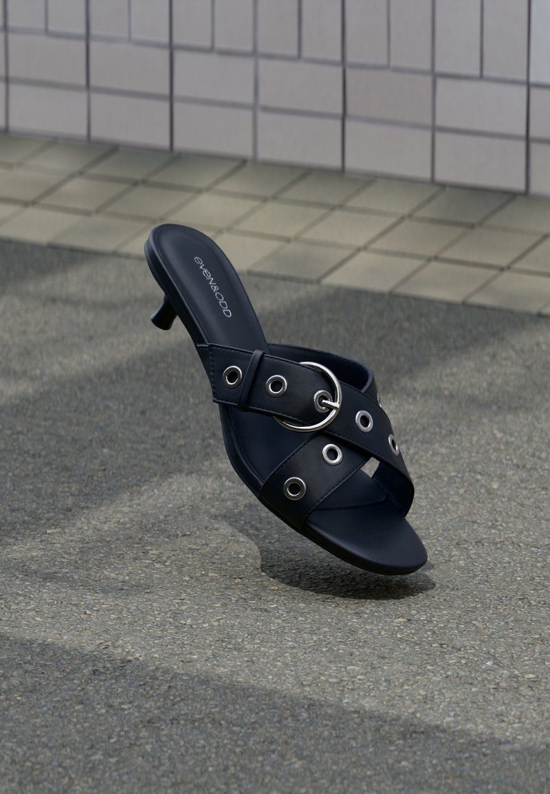 Μαύρο ψηλοτάκουνο σανδάλι με σταυρωτούς ιμάντες που διαθέτουν ασημένιες ροδέλες και στρογγυλό πόμολο, τοποθετημένο σε υφή πλακόστρωτου κοντά στον τοιχο με πλακάκια.