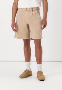Pantalones cortos de algodón beige con pliegues frontales, bolsillos laterales y cierre de botón, combinados con zapatillas marrones y calcetines blancos sobre un fondo liso.