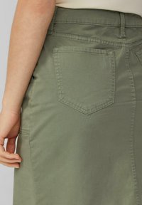 Jupe vert olive avec une texture lisse, présentant une poche arrière et des détails de couture. La ceinture se positionne à la taille pour un look ajusté.