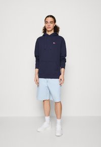 Marineblaue Kapuzenjacke mit einer Fronttasche und Kordelzugkapuze, kombiniert mit hellblauen Jeansshorts und weißen Sneakers.