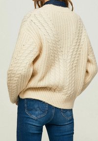 Kremowy sweter z warkoczowym splotem, z szerokimi rękawami i ściągaczem, noszony na ciemnych dżinsach, eksponujący wyraźne wzory oraz teksturę dzianiny.