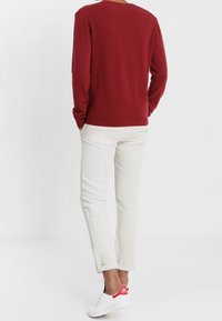 Man som står med händerna i fickorna, klädd i en röd långärmad skjorta, ljusa byxor och vita sneakers med röda detaljer.