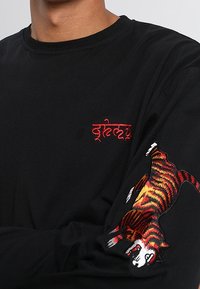 Musta pitkähihaine paita, jossa on punaiset brodeeratut tekstit rinnassa ja värikäs tiikeri-kuvio oikealla hihalla. Pehmeä kangas, istuva malli.