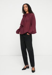 Burgundy, long-sleeve blouse with a tie neck and gathered details, paired with high-waisted black jeans and black slingback shoes.