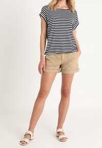 Camisa de rayas azul marino y blanca de manga corta combinada con pantalones cortos caqui de puño enrollado. Sandalias blancas con herrajes decorativos visibles.