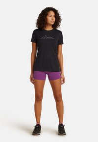 Sort bomulds T-shirt med bjerg- og månegrafik, parret med lilla shorts. Modellen står med sneakers op ad en hvid baggrund.