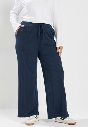 Personne portant un pantalon large bleu marine avec une taille à cordon et des baskets blanches avec des rayures noires.
