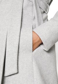 Lichtgrijze wollen jas met een gestructureerde afwerking, zijzakken, een taille met ceintuur en een reverskraag. De stof lijkt zacht en warm.