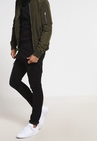 Chaqueta bomber verde oliva con bolsillo con cremallera, camiseta negra de mangas largas, jeans ajustados negros y zapatillas blancas. Diseño simple, acentos mínimos.