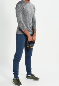 Man in gray long-sleeve shirt and blue pants holding a black kettlebell with a yellow stripe, standing on a white background.